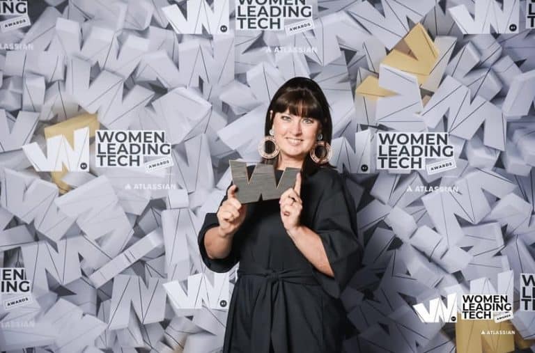 Woman with dark hair standing in front of media wall holding award shaped like the letter W.